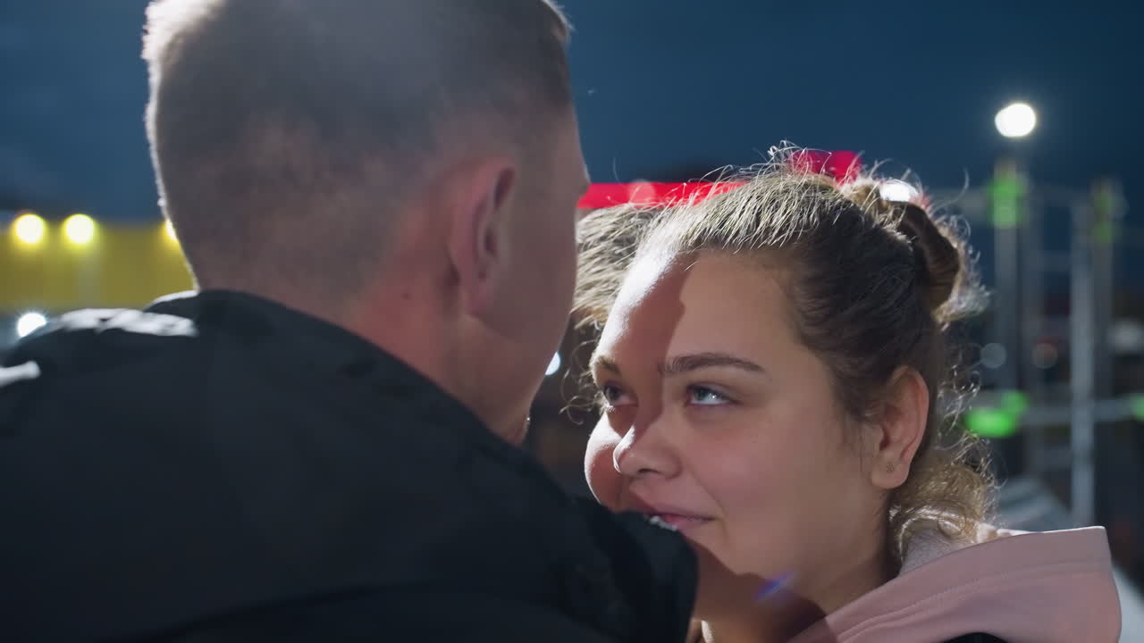Young man kisses his love on forehead under glowing street light as she closes eyes and smiles softly with blurred city background filled with colorful lights