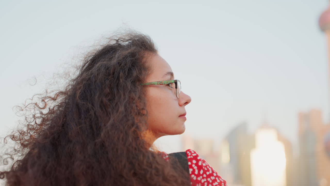 Donna riccia osserva con calma il paesaggio urbano distante. Donna urbana in abito rosso contempla lo skyline sotto un bagliore dorato. Donna multirazziale con gli occhiali guarda pensierosa le torri della città durante il tramonto