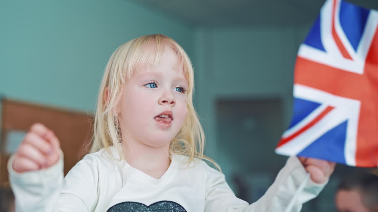 Cute little girl playing with British flag. English flag with three red crosses on blue and white background. Flag of Great Britain in child's hands indoors.