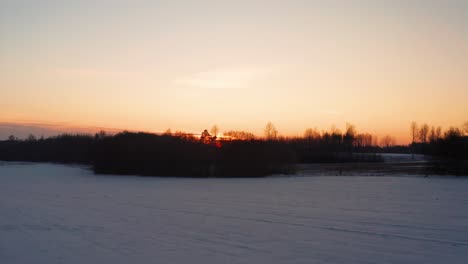 Flying-above-snowy-agricultural-field-toward-sunset-forest-silhouette