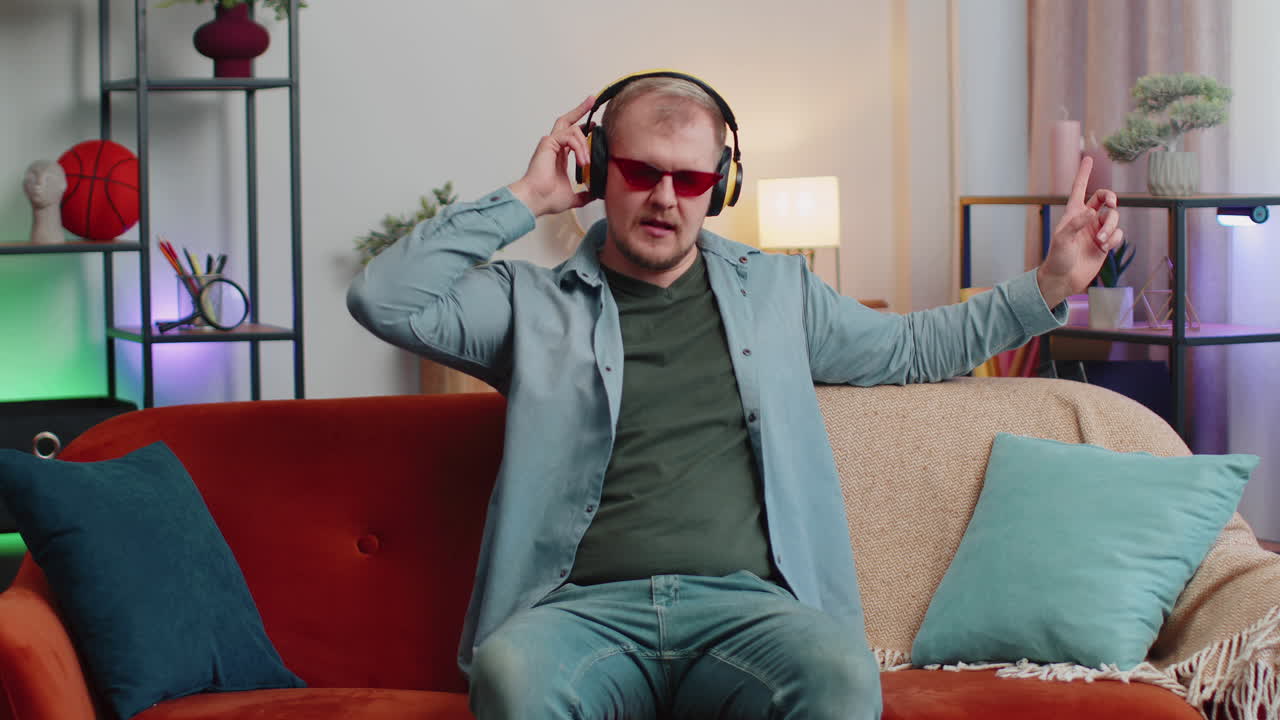 무선 헤드폰을 들고 춤을 추고, 집의 거실에서 편안한 소파에서 노래를 부르는 즐거운 젊은 남자