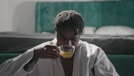 man in white robe sipping tea, eyes focused on cup in tranquil bedroom. soft light from window highlights peaceful atmosphere, creating serene and relaxing moment for self-care and contemplation