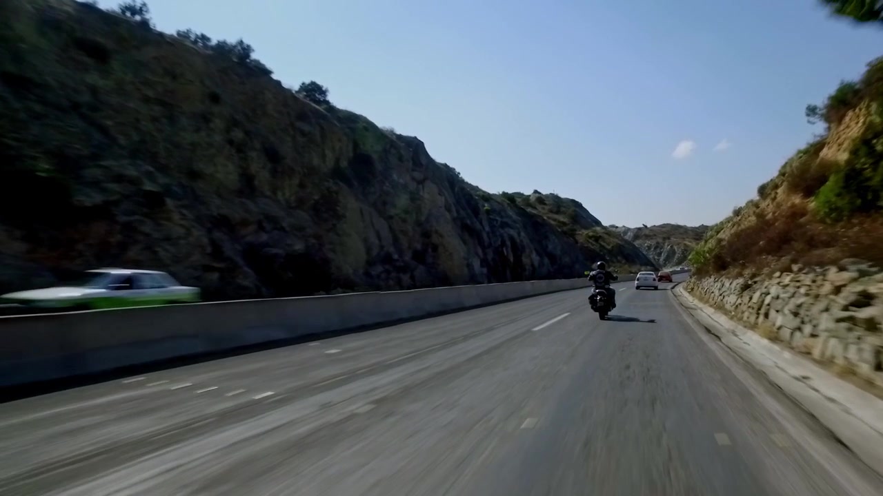 Motociclista che percorre un'autostrada di montagna asfaltata tra scogliere rocciose con veicoli di passaggio. Viaggio panoramico su strada attraverso un paesaggio aspro in una giornata limpida.