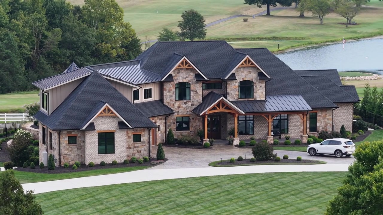 A stone and timber luxury home with a standing seam metal roof overlooks a golf course pond and open fairway on a summer afternoon in Forest, Virginia. Aerial wide shot