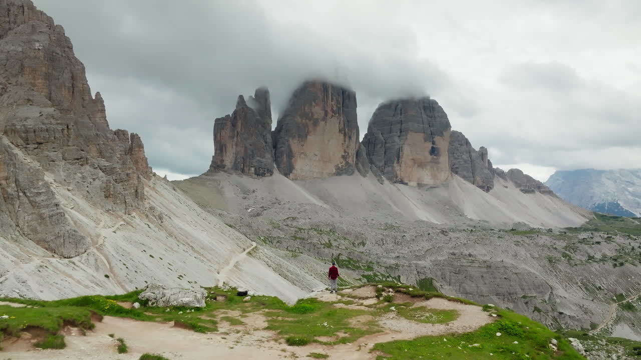 A man standing on edge of mountain overlooking Tre Cime di Lavaredo or Three Peaks limestone face partially covered in clouds, Dolomites, Drone shot