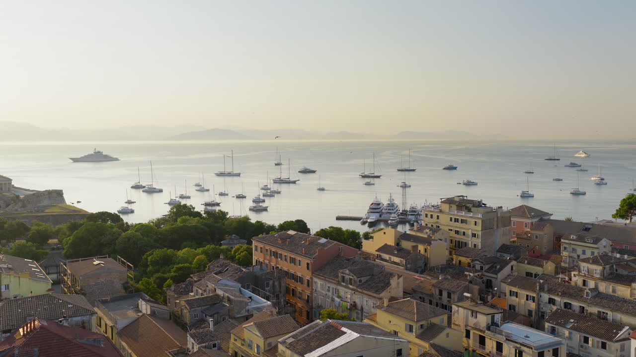 Old Town Corfu at sunrise with harbor, boats, and swallows flying around rooftops, drone push forward to ocean