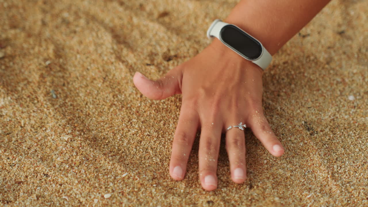 Hand tracing circles on sand beach, closeup of hand with watch and ring smoothing grains into concentric curves, gentle fingertip motion, warm sunlit texture and calming seaside atmosphere for mindful