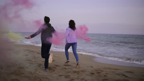 vista posterior de una joven pareja corriendo con una granada de humo de color en sus manos junto al mar durante el atardecer. divertido, humo colorido a su alrededor