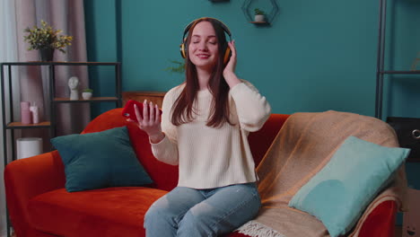 Overjoyed-young-woman-in-wireless-headphones-dancing,-singing-on-cozy-couch-in-living-room-at-home