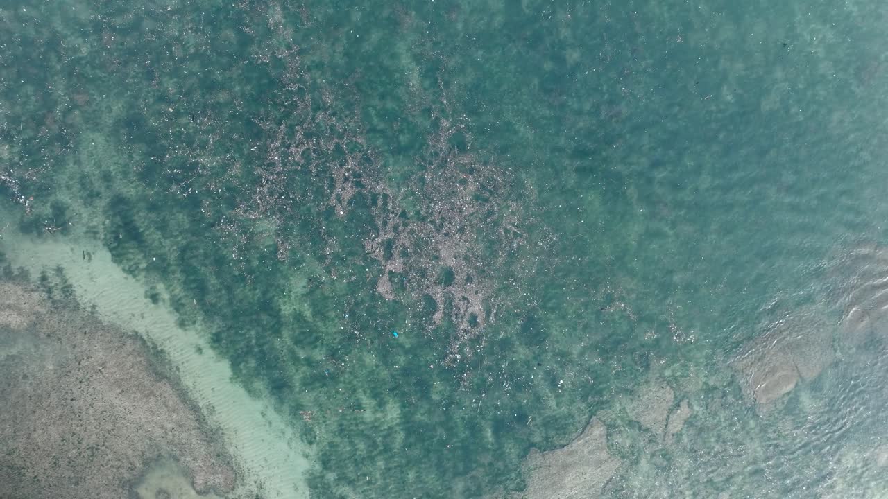 High altitude top down ascending drone shot of polluted water filled with trash floating over dead coral reef in the turqouise tropical water of Bali Indonesia