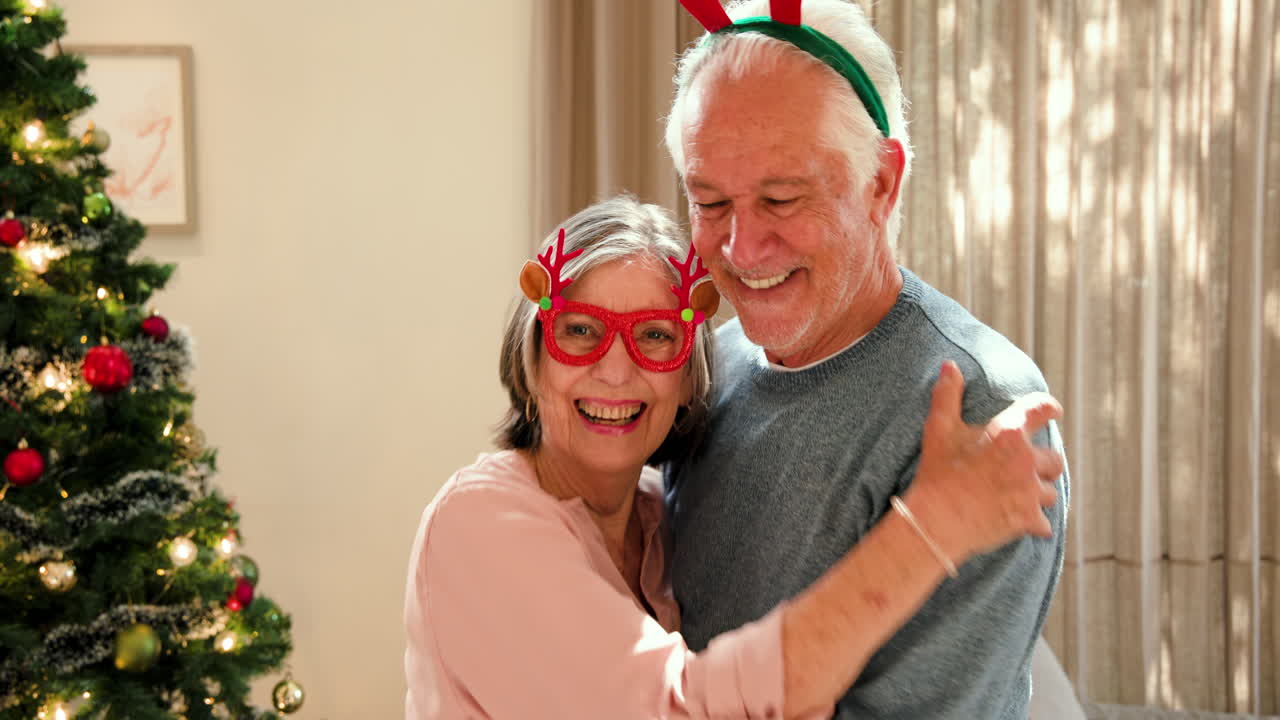 Senior couple wearing festive accessories smiling joyfully by Christmas tree at home