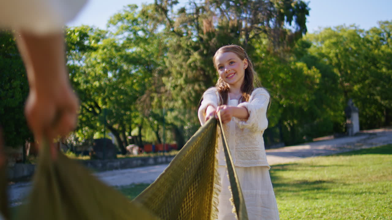 Happy girl preparing picnic helping mother spread out blanket at summer grass
