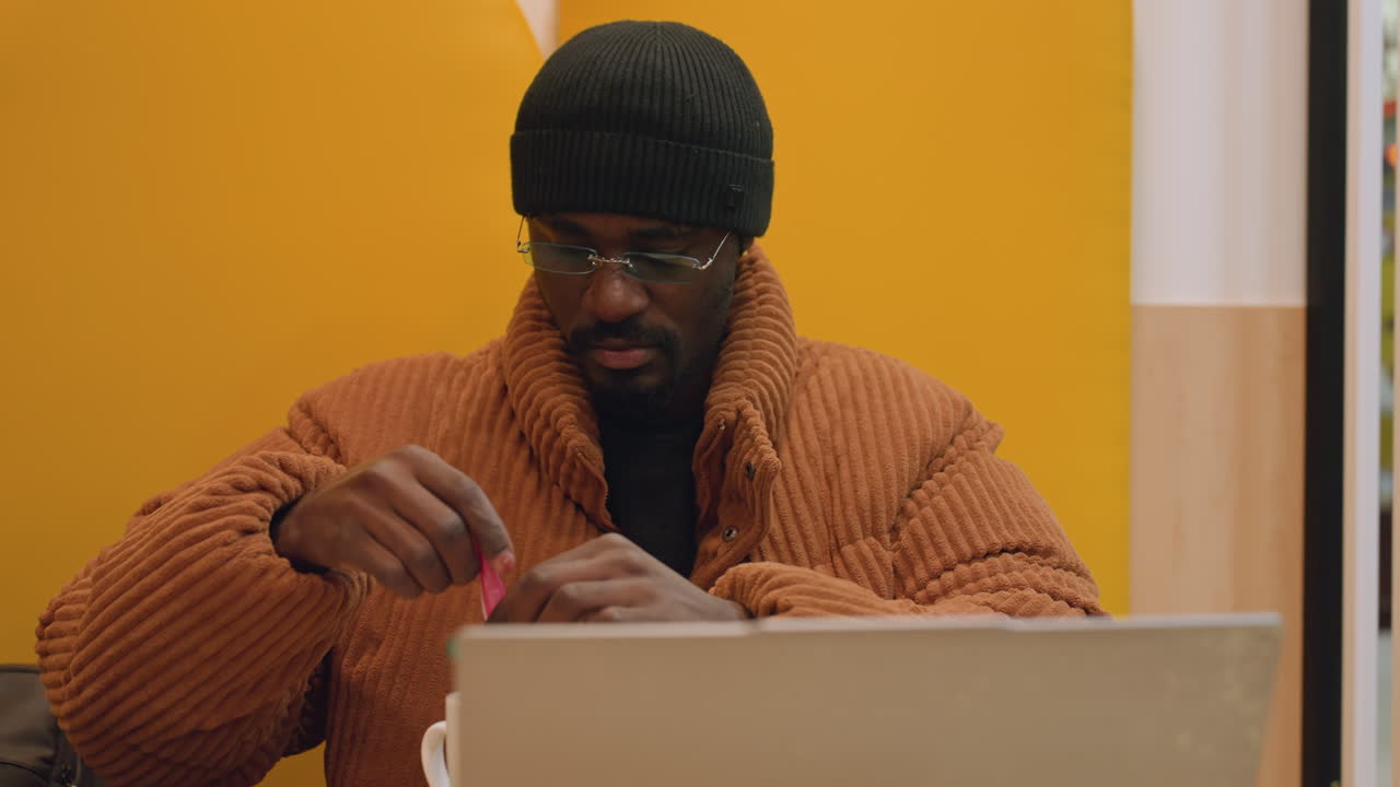 Black man opening small snack during break in yellow booth, unwraps candy near laptop and coffee cup, focused on hands with calm expression, casual coat and beanie, soft mall ambience and relaxed