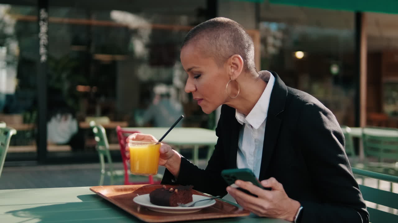 Woman with Shaved Head Using Phone and Drinking Juice at Outdoor Cafe