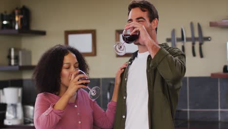 Happy-diverse-couple-standing-in-kitchen,-drinking-red-wine-and-embracing