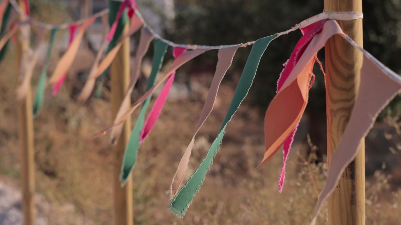 Bandiere di tessuto vivaci in verde, arancione, rosa e beige sventolano con grazia al rallentatore su pali di legno sotto una morbida luce solare naturale, creando una scena festosa all'aperto senza persone presenti
