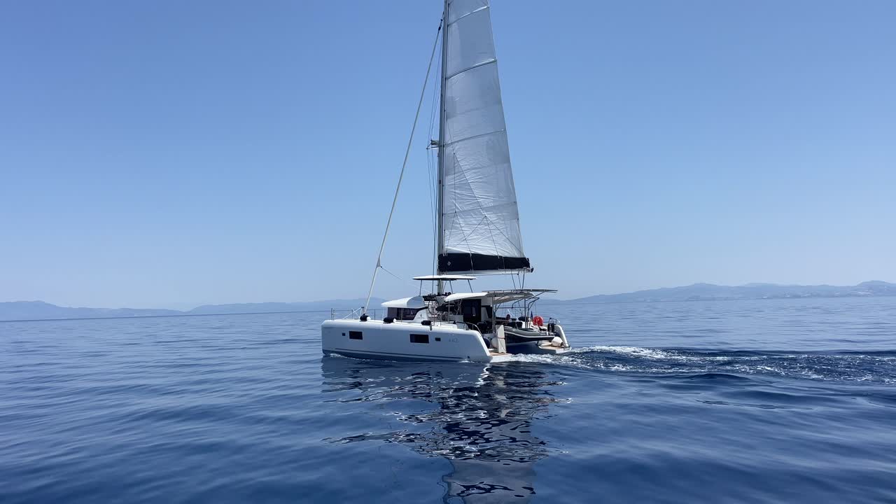 Greece, Samos Island, a white catamaran with its sail is on, moving slowly on a sunny day with blue skies, no clouds and calm sea.