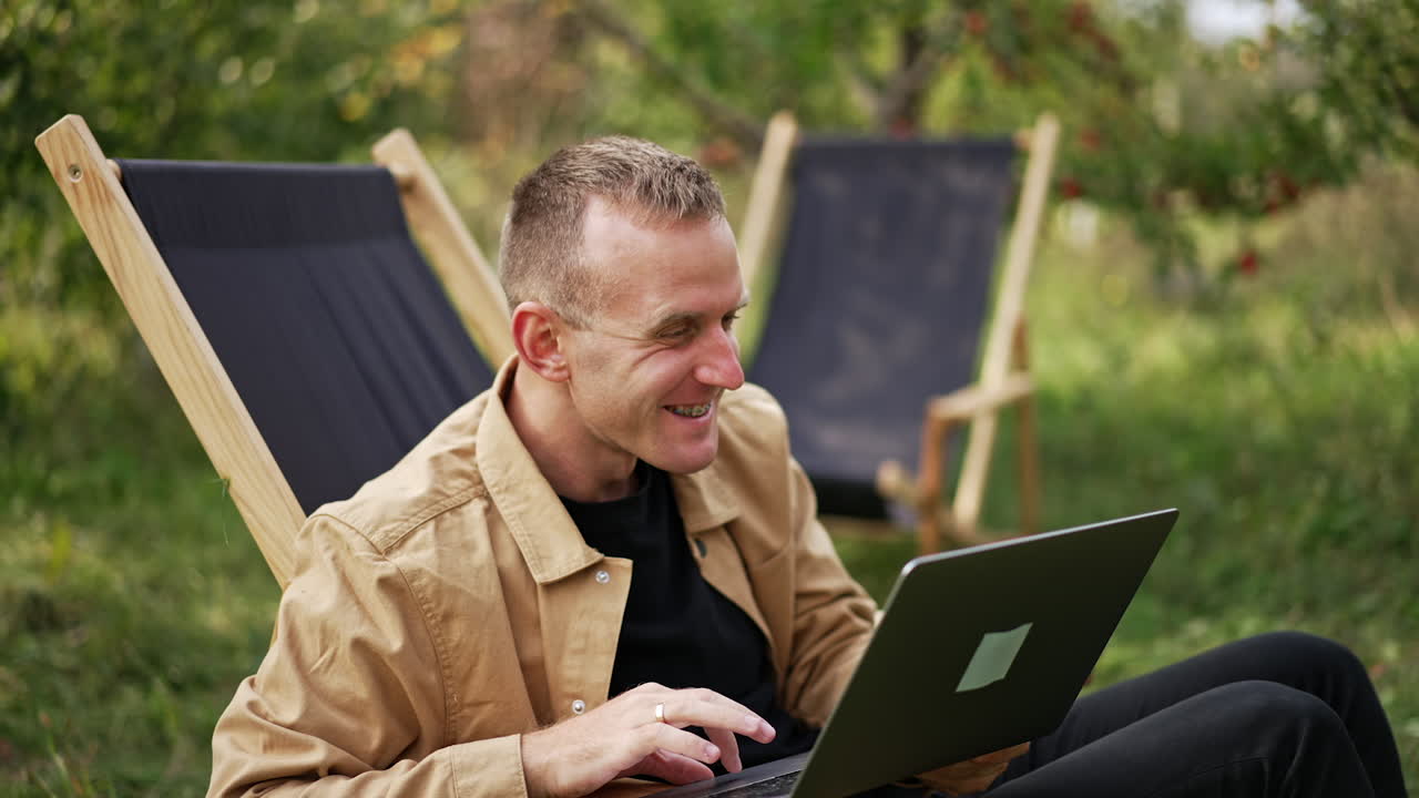 Mid aged man working on his laptop sitting in the garden chair outside. Happy smiling Caucasian freelance male using internet outdoors on computer.