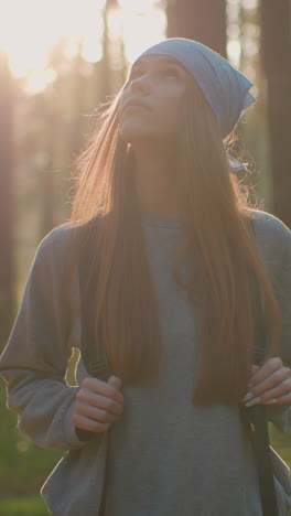 una mujer joven con una mochila camina a través de un bosque sereno, suavemente iluminado por la cálida luz del sol de la tarde por detrás, con una mirada reflexiva mientras camina