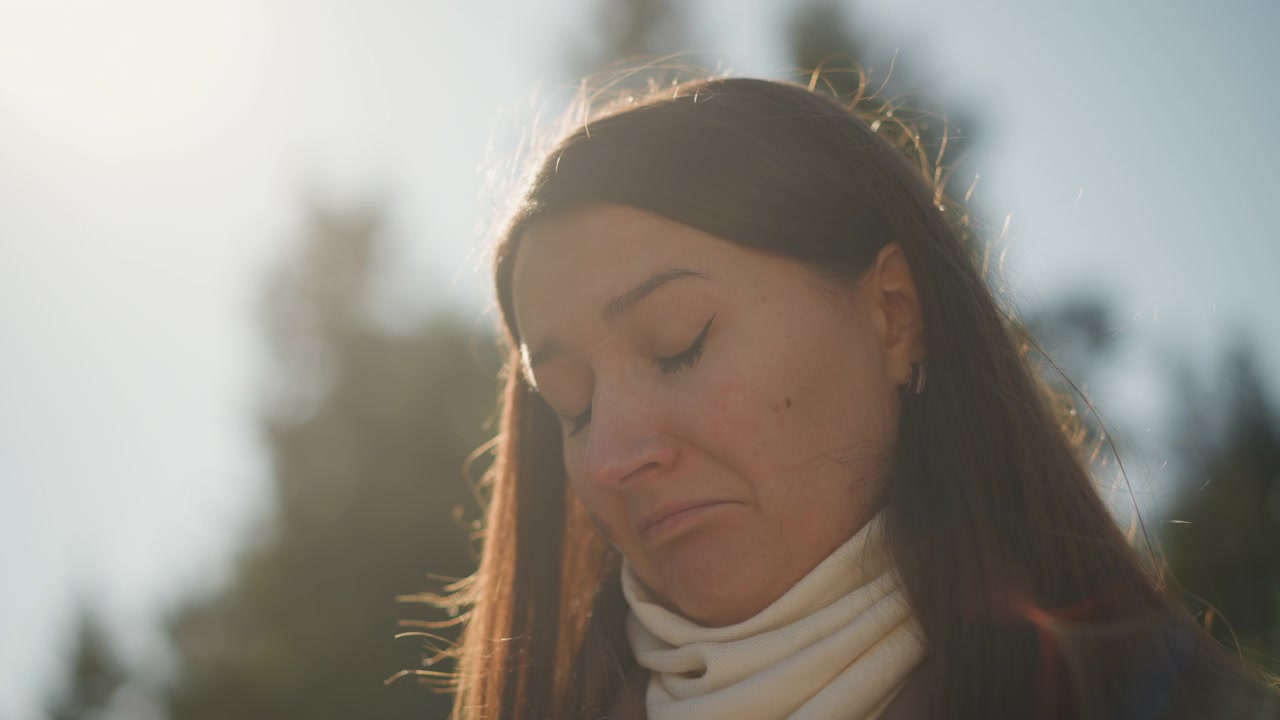 Plano detalhe de mulher com cabelo castanho longo, vestindo gola alta branca, em pé ao ar livre sob luz solar suave. Sua expressão é calma e introspectiva enquanto ela olha para baixo contra um fundo desfocado.