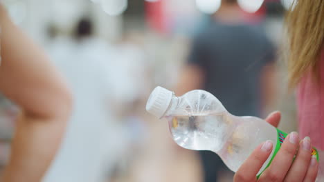 lady holds a chilled bottled water in a mall behind another person, with a well-lit, blurred background featuring other people walking around, the bottle is clear and cold with moisture on it