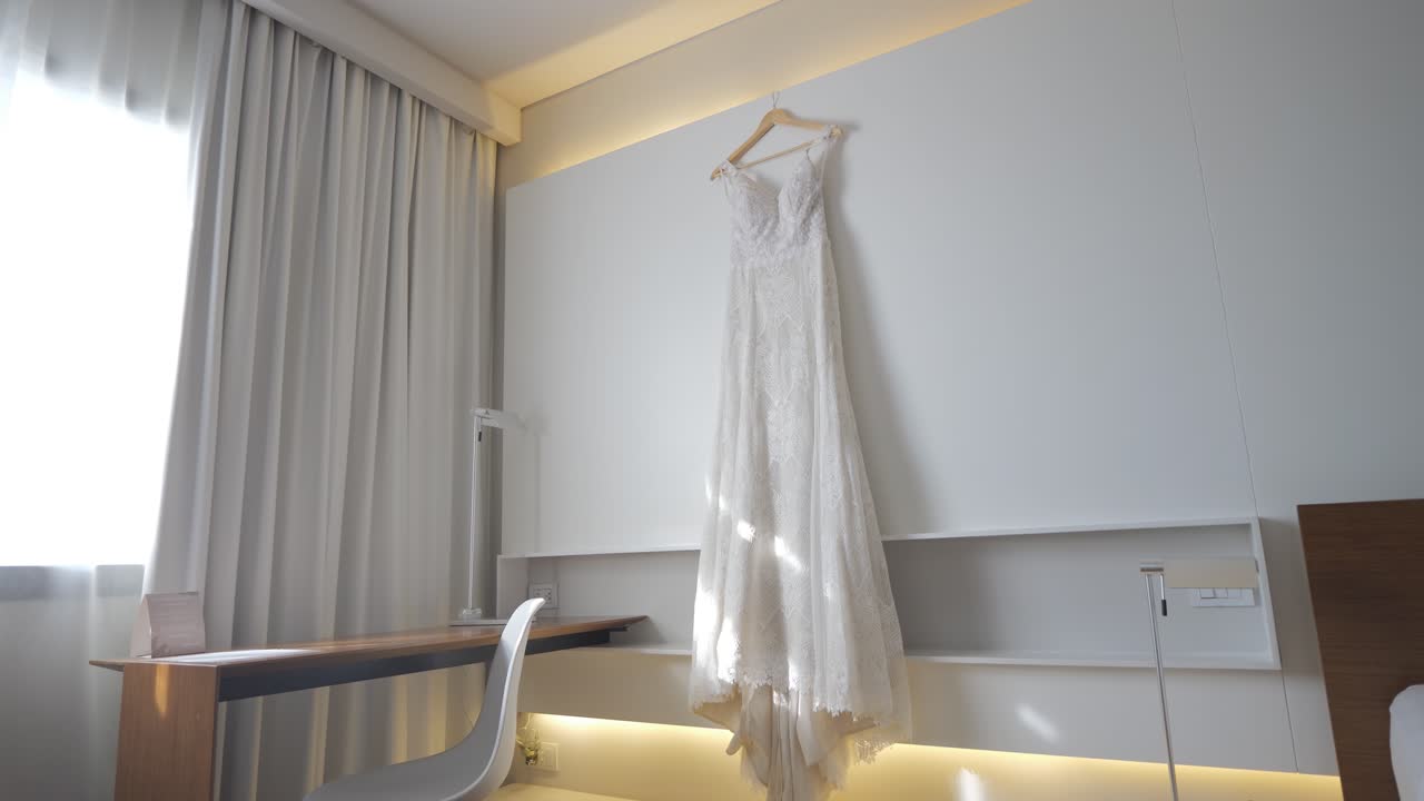 A white dress is hanging on a clothesline in a hotel room. The dress is long and flowing, and it is hanging from a clothesline on the wall. The room is clean and well-lit, with a desk