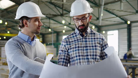 two caucasian men wearing helmets talking and looking at a blueprint in a factory