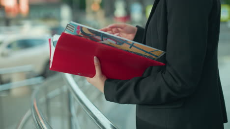 side view of woman in black blazer examining vibrant artistry book in red folder while standing near iron railing in outdoor urban area with soft blur of traffic signs and busy city background