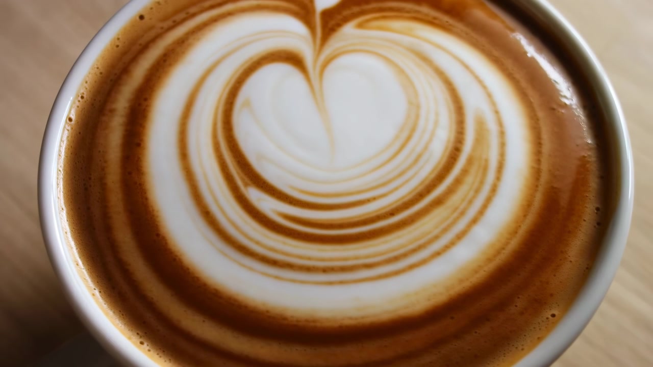 Una vista ravvicinata dall'alto segue un movimento sottile su una superficie di caffè appena decorata, con schiuma cremosa a forma di cuore, linee marroni concentriche, il bordo della tazza in ceramica e un caldo tavolo di legno visibili.