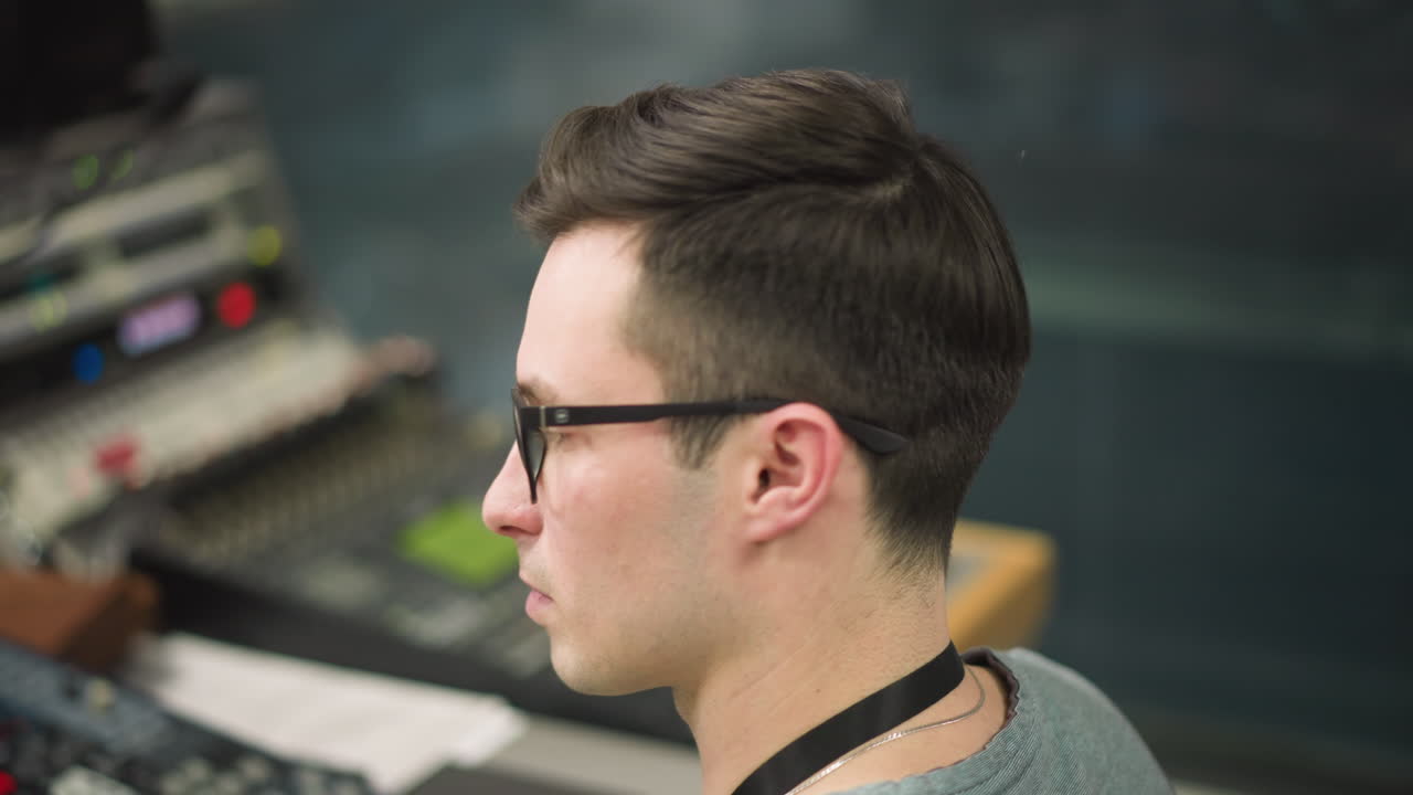 Young man with glasses focused on computer screen in office environment, working on digital content or broadcast tasks, close-up shot in modern studio or control room