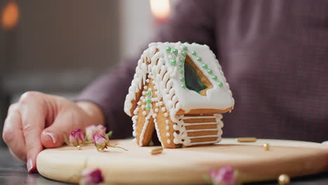 close-up of baker carefully rotating decorated gingerbread house with white icing and green beads, wooden tray, golden sprinkles, dried flowers, and blurred candlelight in background