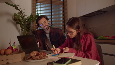 A-confident-and-hardworking-brunette-girl-with-glasses-in-a-red-sweater-studies-foreign-languages-and-writes-notes-in-her-notebook-together-with-a-young-brunette-male-teacher-in-a-modern-apartment-in-the-kitchen