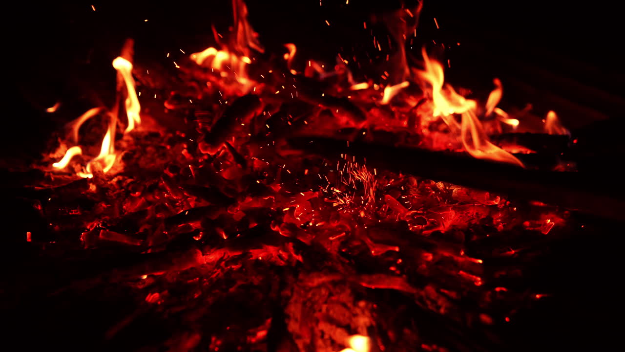 Burning out pieces of wood in the campfire. Unknown person moves the charcoal and sparkles fly up. Close up. Darkness at backdrop.