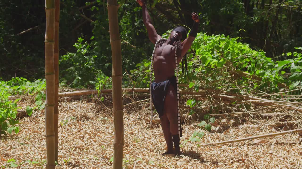 A dark-skinned, muscular man, sporting a Tarzan cosplay outfit, poses within a dense forest.
