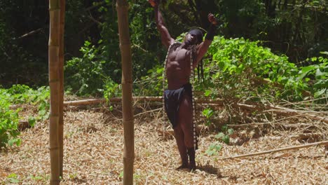 a dark-skinned, muscular man, sporting a tarzan cosplay outfit, poses within a dense forest.