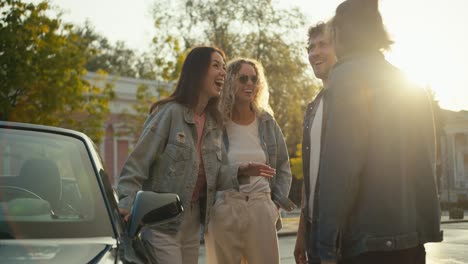 a group of young people are standing and talking on the street near the car. guys are stylishly dressed and having fun against