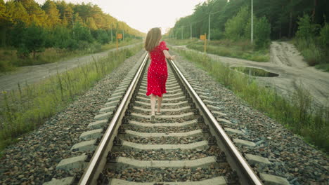 rear view of woman in red dress running along railway track in countryside with surrounding greenery and forest trees during sunset, hair flowing freely as she moves with energy and grace