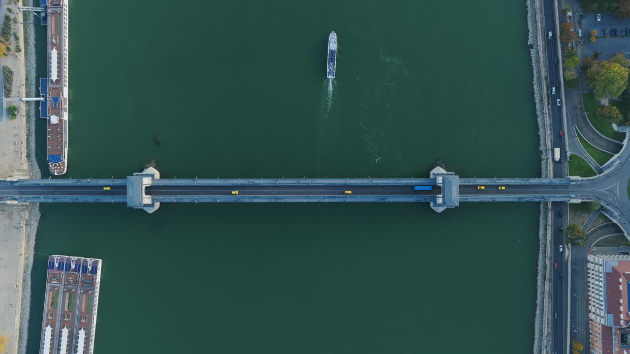 Top-down aerial showing a ship cruising under a central bridge with traffic and moored riverboats