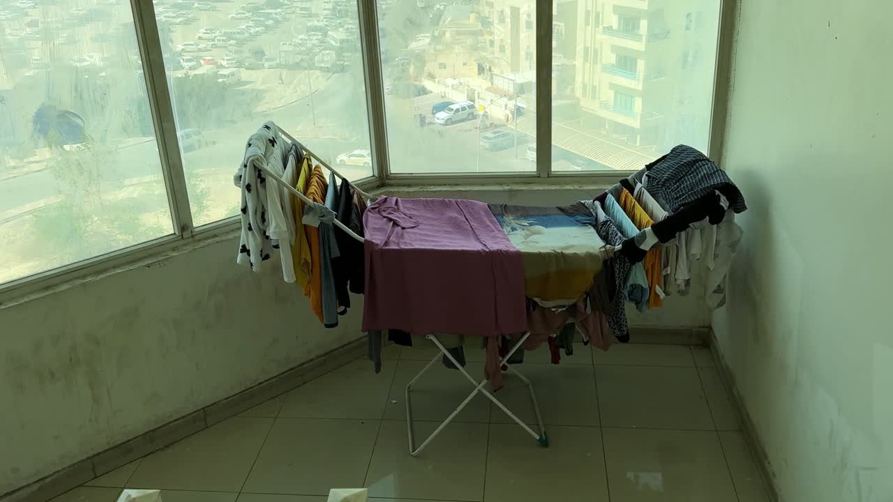 Wide indoor shot of a foldable drying rack filled with clothes near large apartment windows overlooking a city street. Daylight urban view highlights everyday home laundry routine
