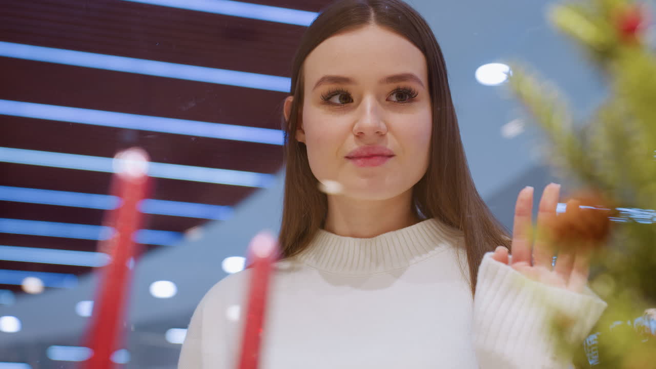 Lady admiring festive decor through glass in a stylish shopping mall with beautiful interior, highlighting modern design, lights, and holiday ambiance