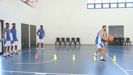 multiracial male basketball players bouncing basketball with coach giving instructions in gym