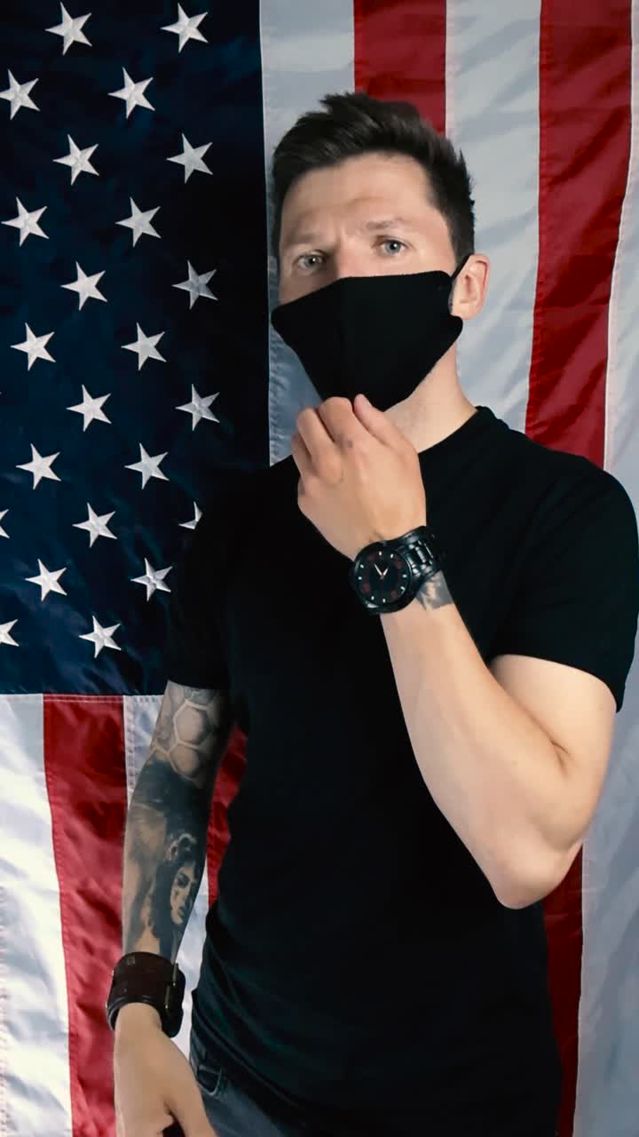 Tattooed white man who is standing in front of a vertical American US flag shows how to properly wear a black facial protection health mask. Nose and face covered and nose out, hand motions included