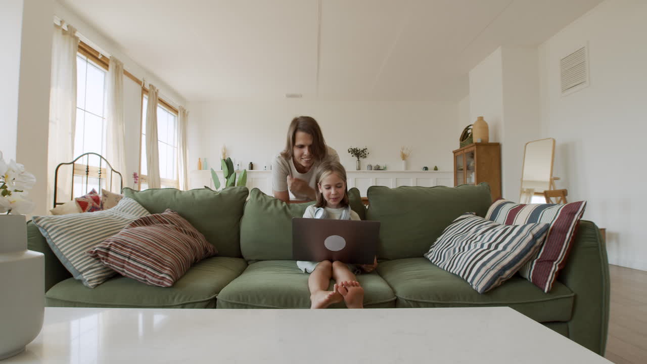 plano general de una niña rubia haciendo su tarea con la computadora portátil y su madre detrás de ella ayudando con la tarea