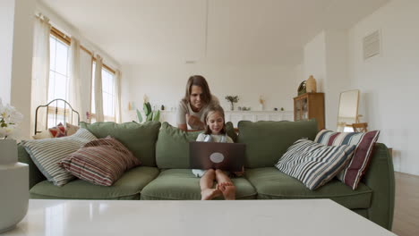 wide shot of a blonde little girl doing her homework with the laptop and her mother behind her helping with the task