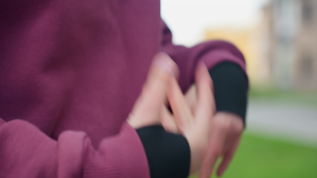 Hoodie clad gym goer presses palms together and slowly flexes fingers in mindful hand stretch to release tension before beginning outdoor exercise under open sky amidst urban fitness park
