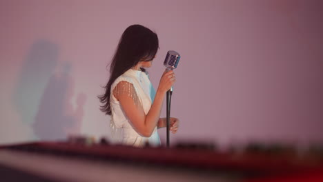 a female singer wearing a white gown stands in front of a vintage microphone, holding it with one hand while moving her head slowly. the background is grey