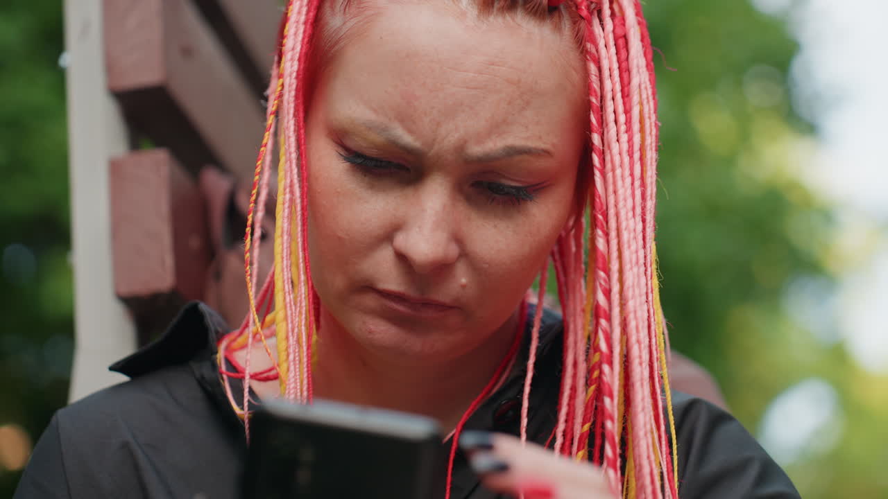 Белая женщина трет лоб, глядя в телефон; неоновые косички и маникюр обрамляют обеспокоенное лицо, напряженное выражение лица и момент личной досады на скамейке, неподдельные откровенные эмоции в свете парка