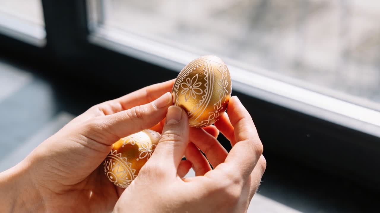 Entering hands holding two golden eggs at window sill, rotating egg to reveal floral pattern
