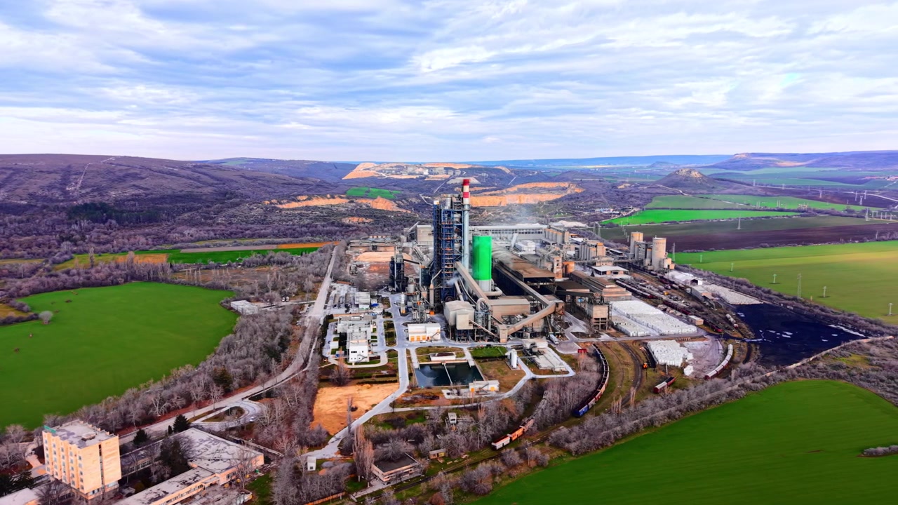 Wide aerial view of cement plant and valley with fields. Drone panoramic of large industrial facility with chimneys and kilns surrounded by green meadows, bare trees, hills and overcast sky.