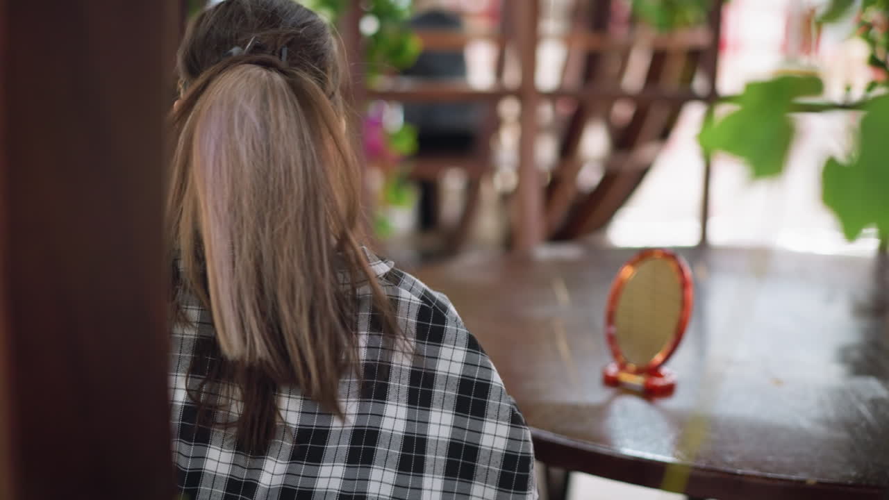 Back view of young lady in checkered shirt placing wooden mirror on table, gazing at her reflection while adjusting hair. Seated in cozy indoor setting with warm lighting